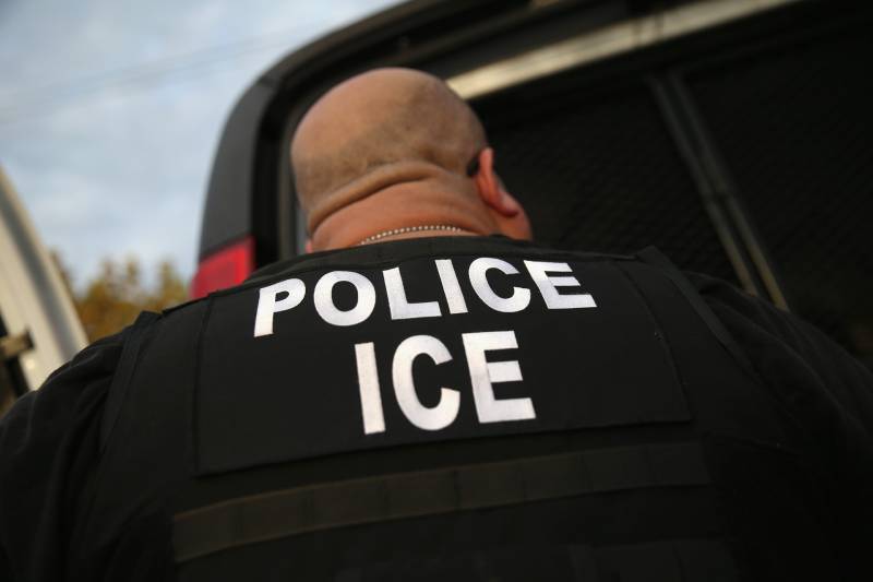Detail shot of the back of a large man with a bald head and thick neck, wearing a black shirt with 'police ice' printed on it in all capital letters