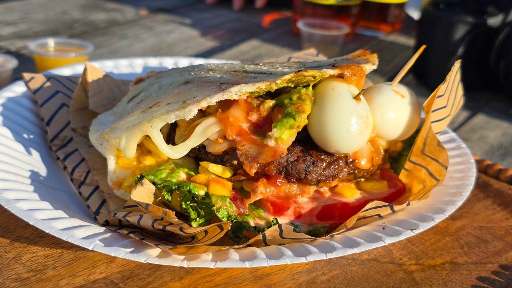An overstuffed arepa, filled with beef patty, quail eggs and more, served on a paper plate.